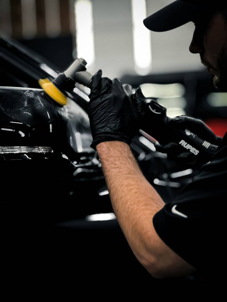 een close-up van een meneer die een zwarte auto aan het polijsten is met zwarte handschoenen en een polijst machine